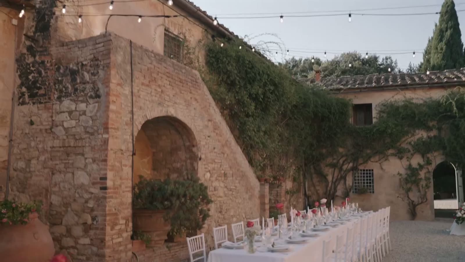 A long dinner table under festoon lights beside the stone walls of the Tuscan villa