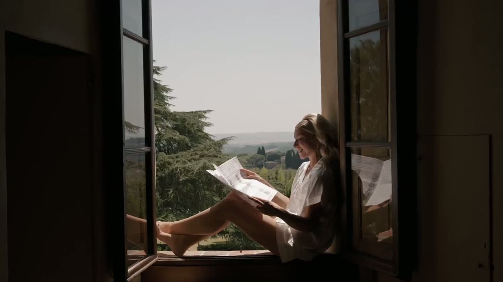 The bride reading the printed wedding gazette on a villa window sill, Tuscan hills beyond